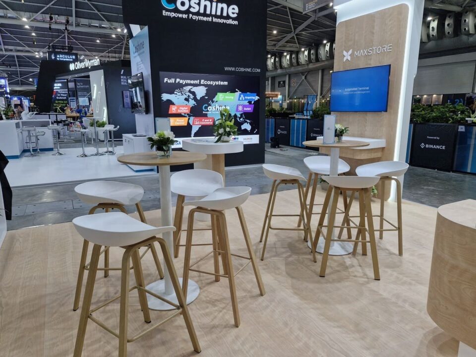 Modern exhibition booth setup with light wood bar stools and round tables, styled for networking and casual seating in a trade show environment.